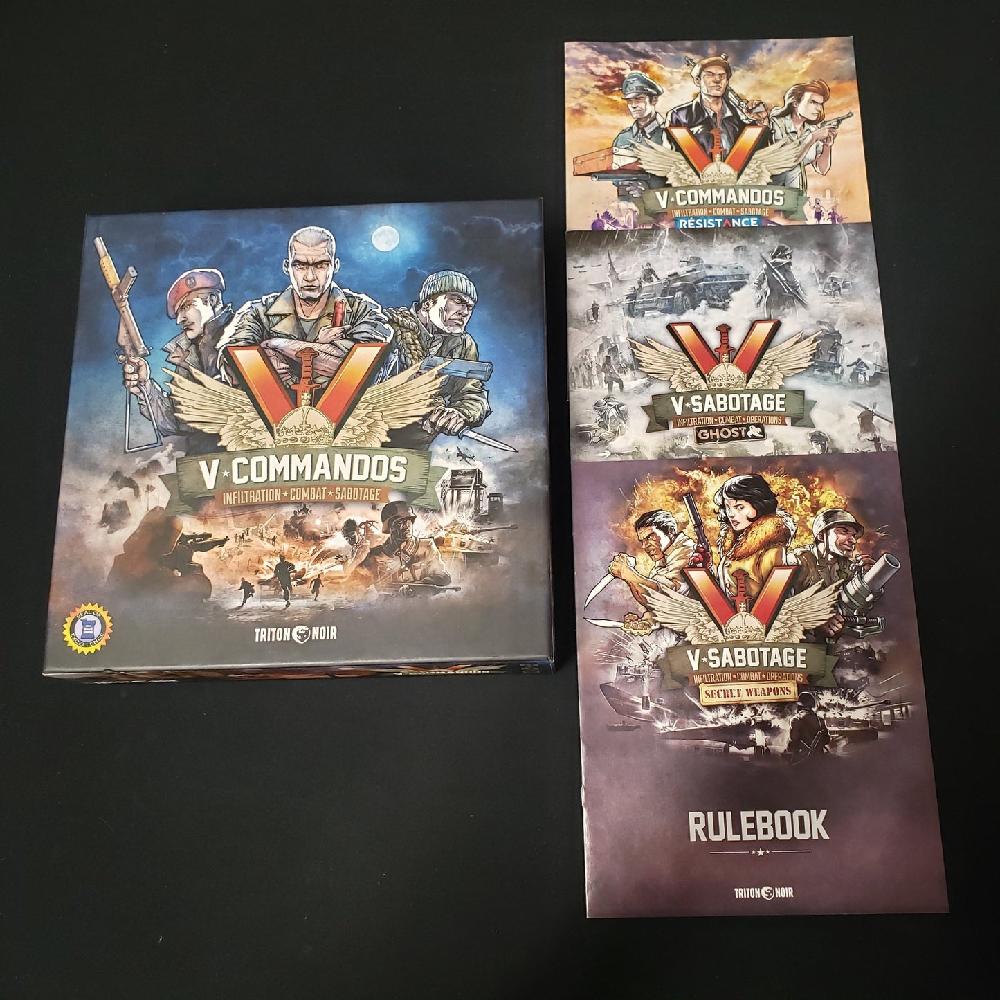 Image shows the front cover of the box of the V-Commandos board game, sitting next to instruction manuals for 3 expansions