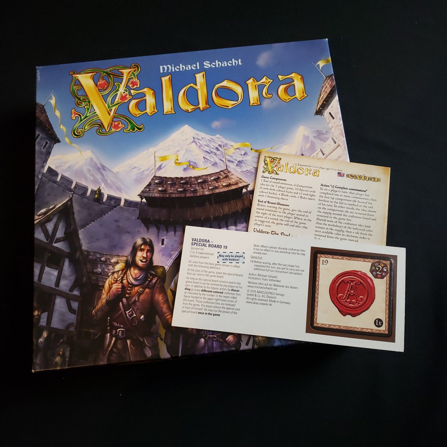 Image shows the front cover of the box of the Valdora board game, with expansion instructions and a promo tile on top of it