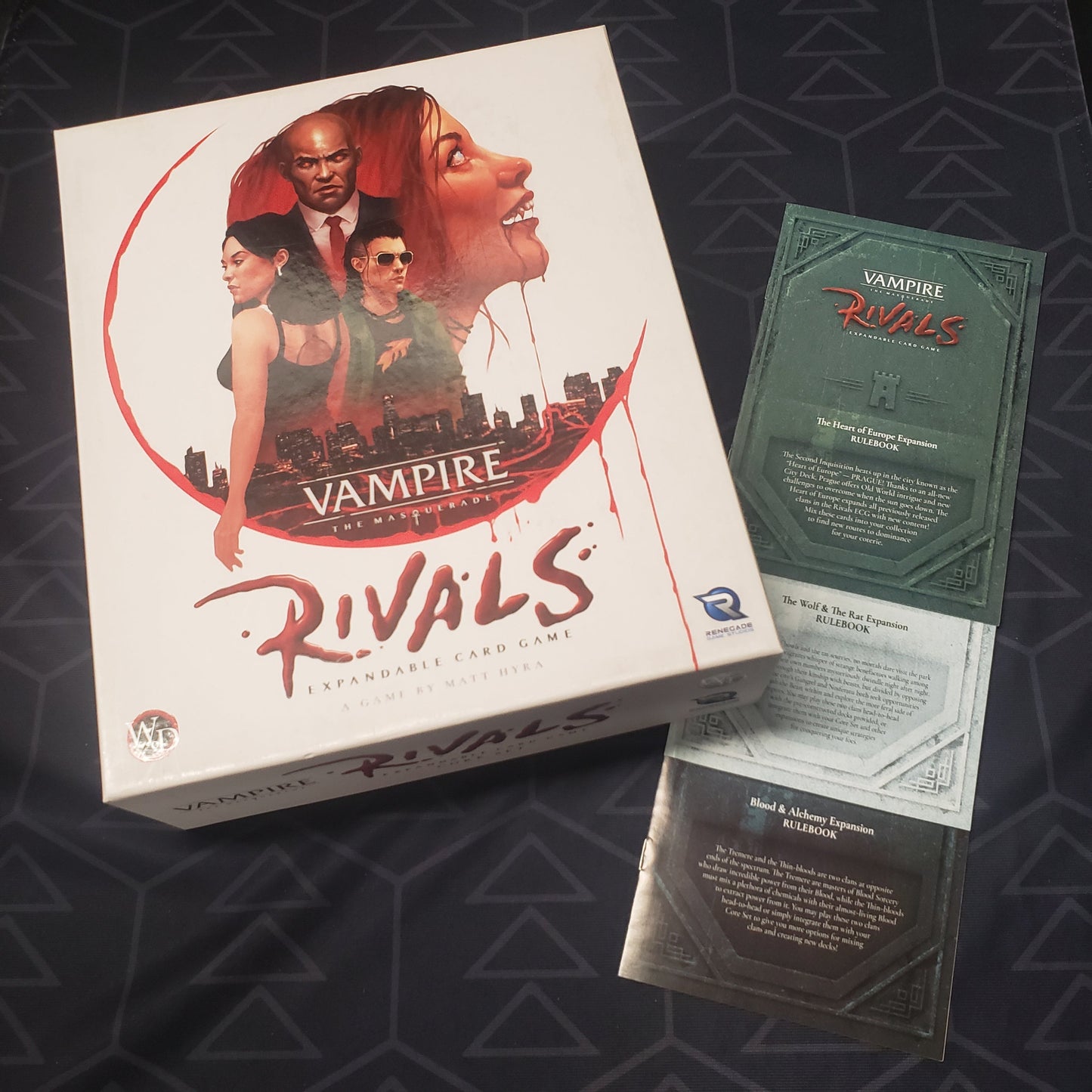 Image shows the front cover of the box of the core set for the card game Vampire The Masquerade: Rivals alongside the instruction booklets for three expansions