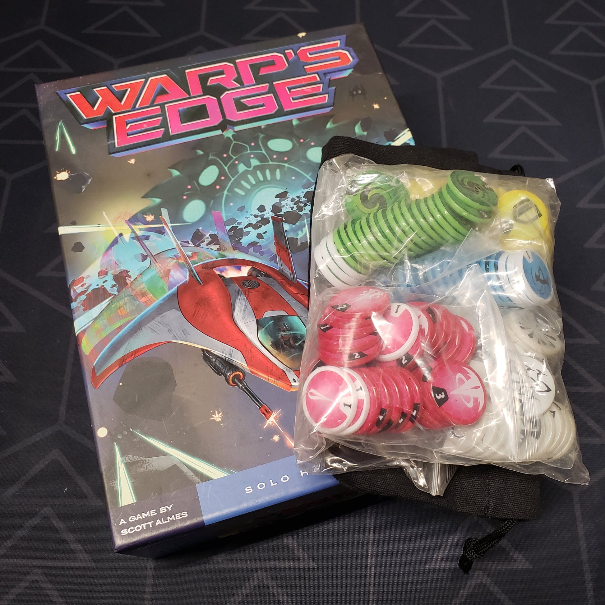 Image shows the front cover of the box of the Warp's Edge board game with a bag of upgraded tokens sitting on top of it