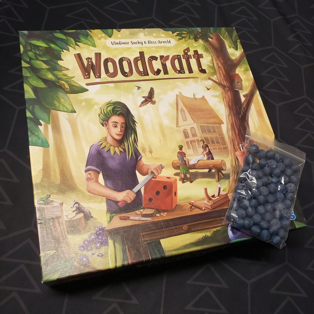 Image shows the front cover of the box of the Woodcraft board game with some upgraded tokens sitting on top of it