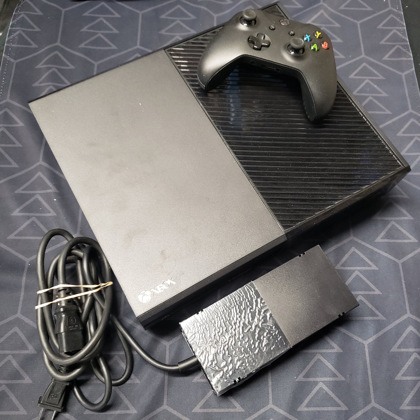 Image shows the top of an Xbox One gaming console with a controller and the power supply