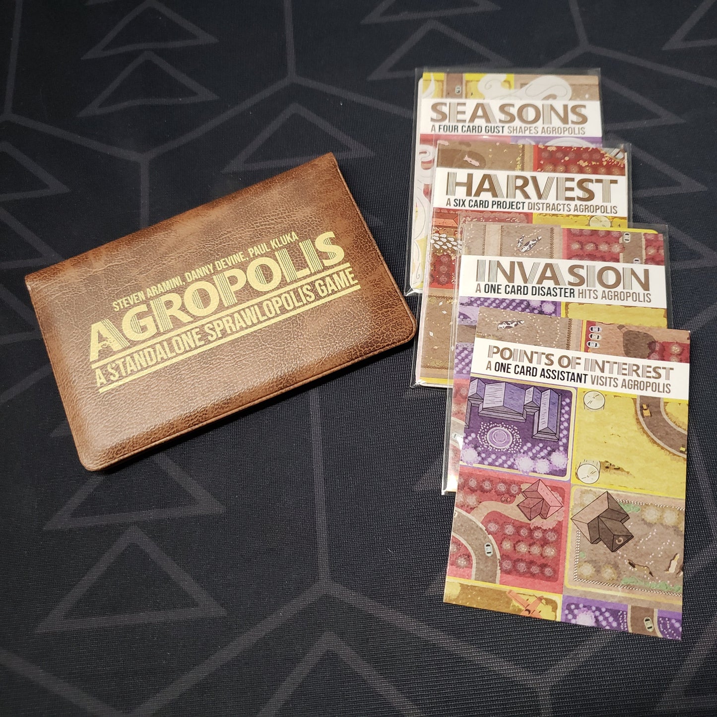Image shows the front of the card wallet for the Agropolis card game, with the Invasion, Points of Interest, Harvest & Seasons expansions sitting next to it