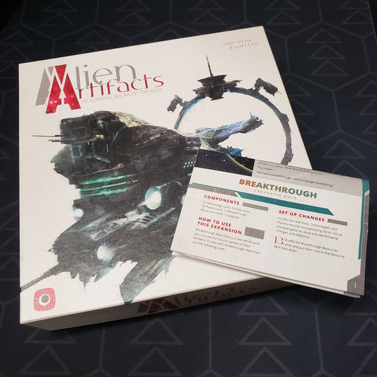 Image shows the front cover of the box of the Alien Artifacts board game with the instructions for the Breakthrough expansion sitting on top of it