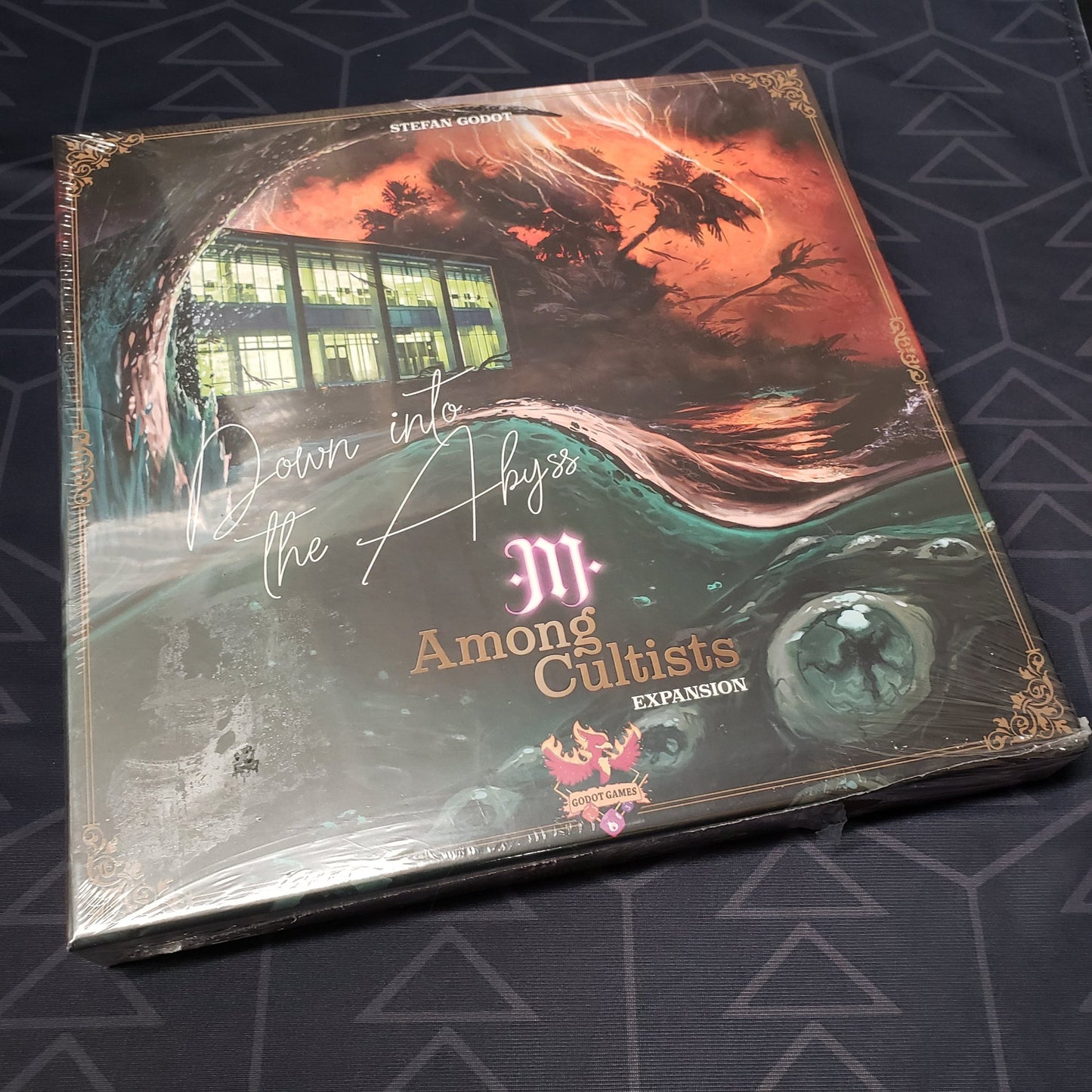 Image shows the front cover of the box of the Down into the Abyss expansion for the board game Among Cultists