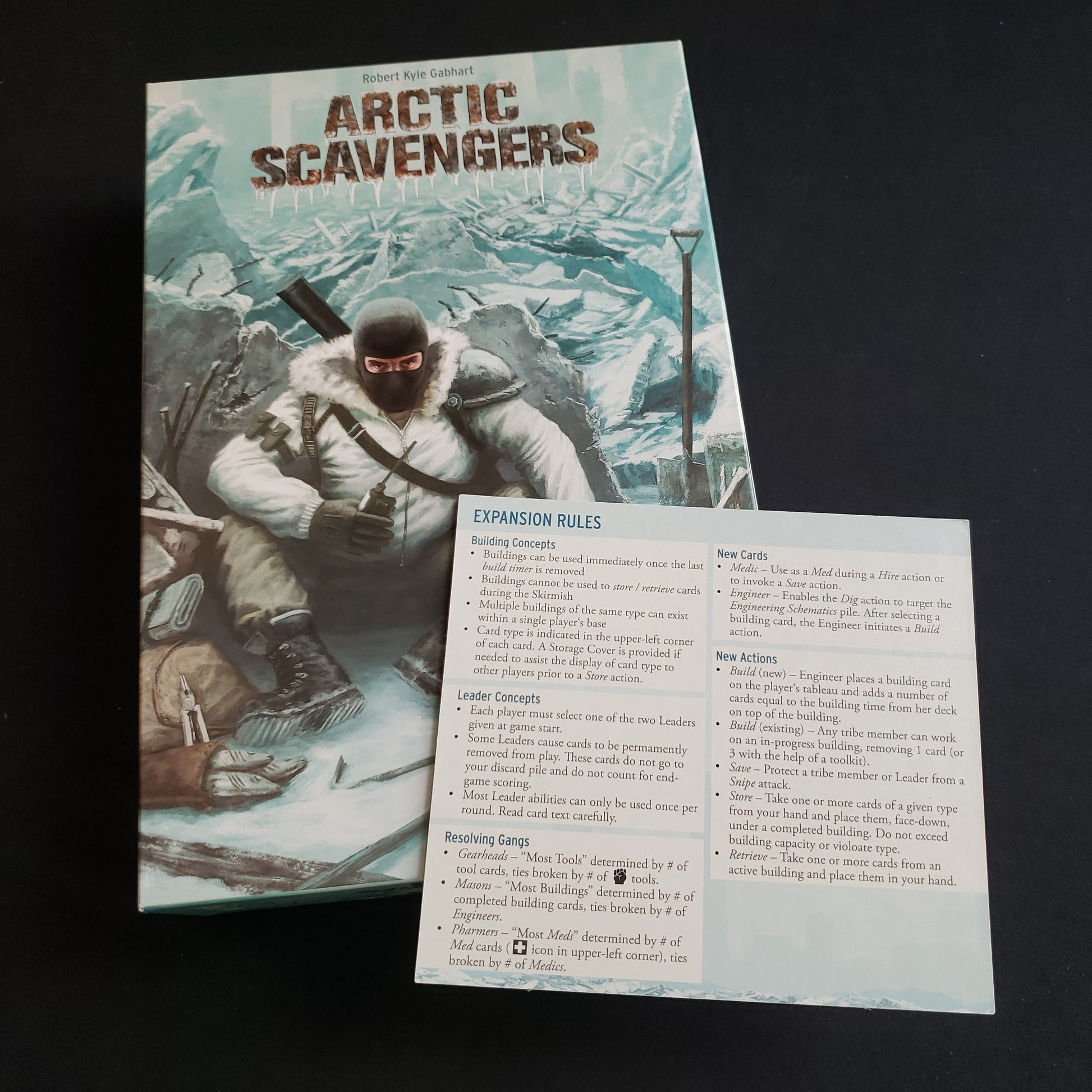 Image shows the front cover of the box of the Arctic Scavengers card game, wit the expansion instructions sitting on top of it