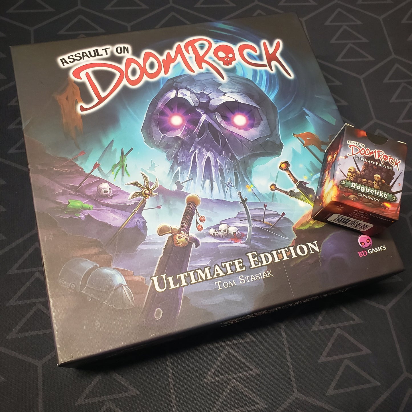 Image shows the front cover of the box for the Assault on Doomrock: Ultimate Edition board game with the Roguelike expansion box sitting on top of it