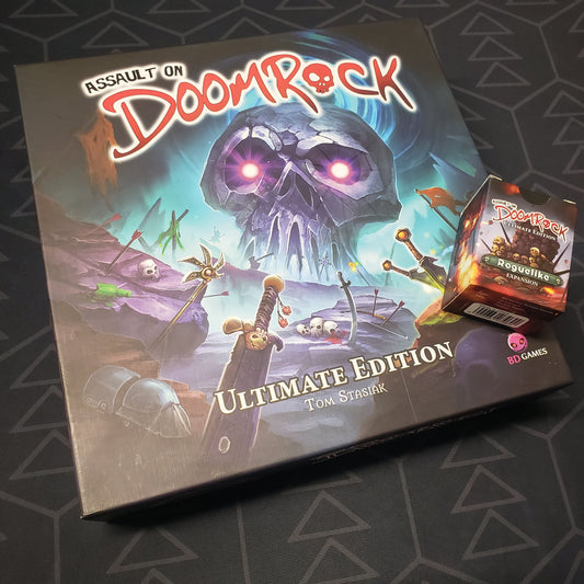 Image shows the front cover of the box for the Assault on Doomrock: Ultimate Edition board game with the Roguelike expansion box sitting on top of it