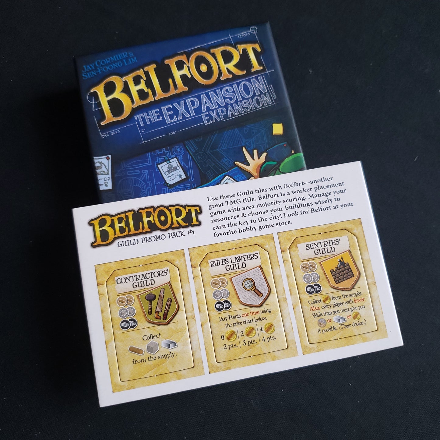 Image shows the front cover of the box of the Expansion expansion for the board game Belfort, along with the unpunched Guild Promo Pack #1 sitting on top of it