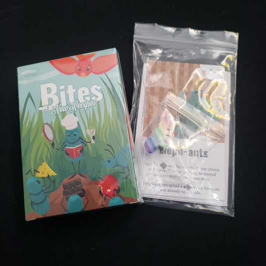 Image shows the front cover of the box of the New Recipes expansion for the card game Bites, with a bagged promo pack sitting next to it