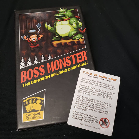 Image shows the front cover of the box of the Boss Monster card game with the instruction card for the Tools of Hero-Kind expansion on top of it