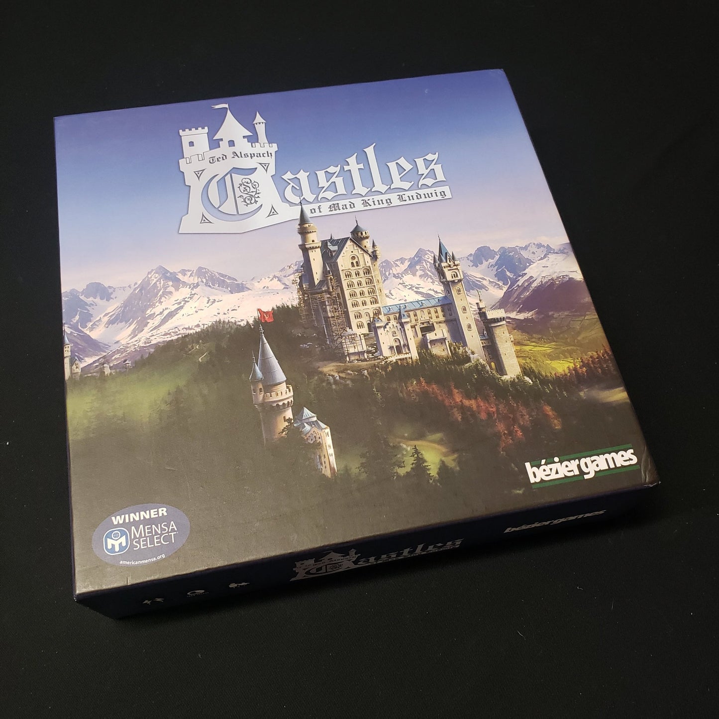 Image shows the front cover of the box of the Castles of Mad King Ludwig board game, with the instructions for the Secrets expansion on top of it