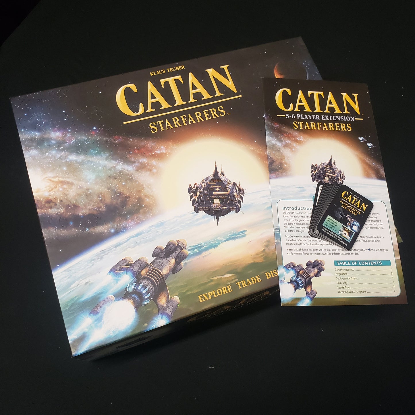 Image shows the front cover of the box for the Catan: Starfarers (Second Edition) board game along with the instructions for the 5-6 lpayer extension and a promo pack of cards