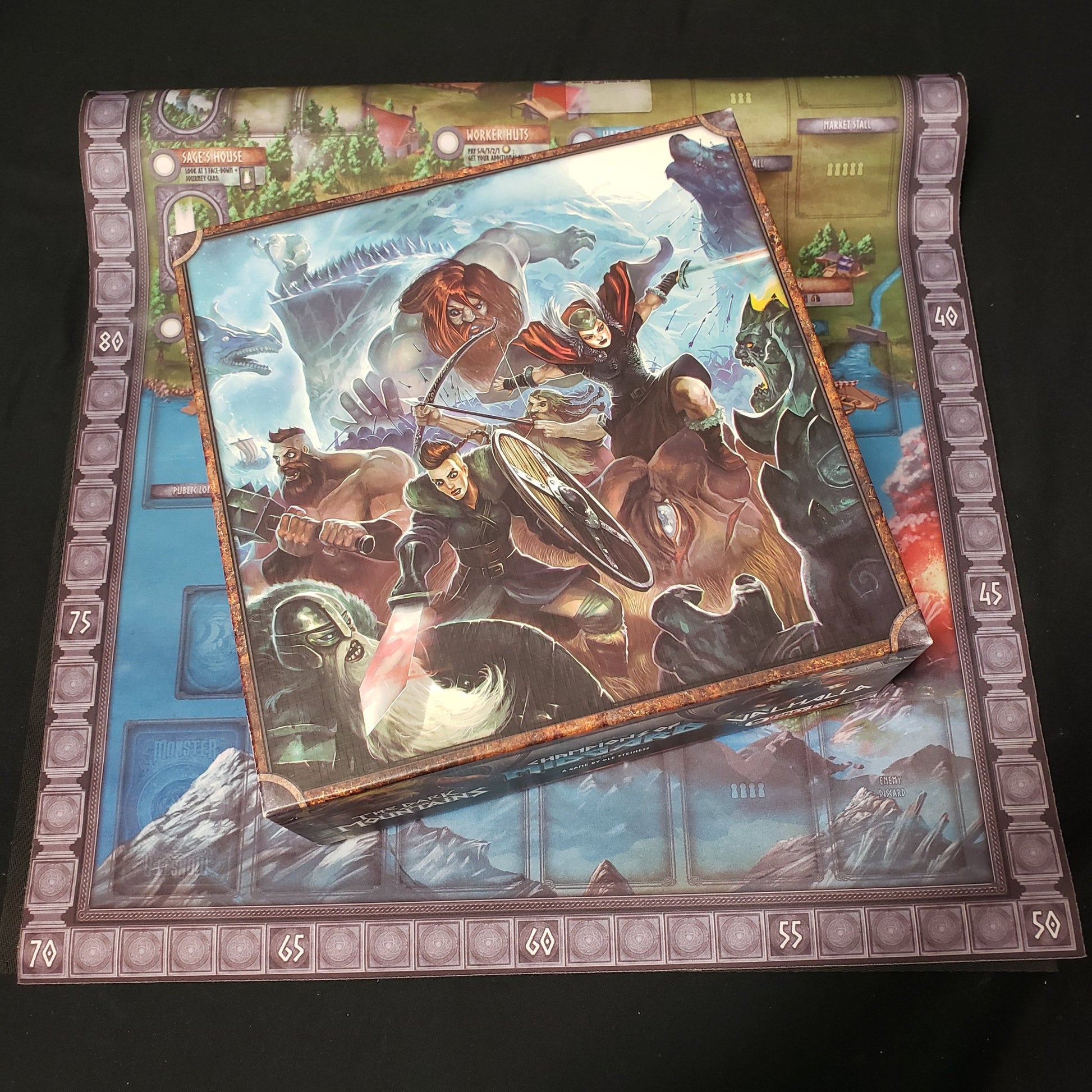 Image shows the front cover of the box of the Jarl Edition of the board game Champions of Midgard, sitting on top of the neoprene playmat for the game