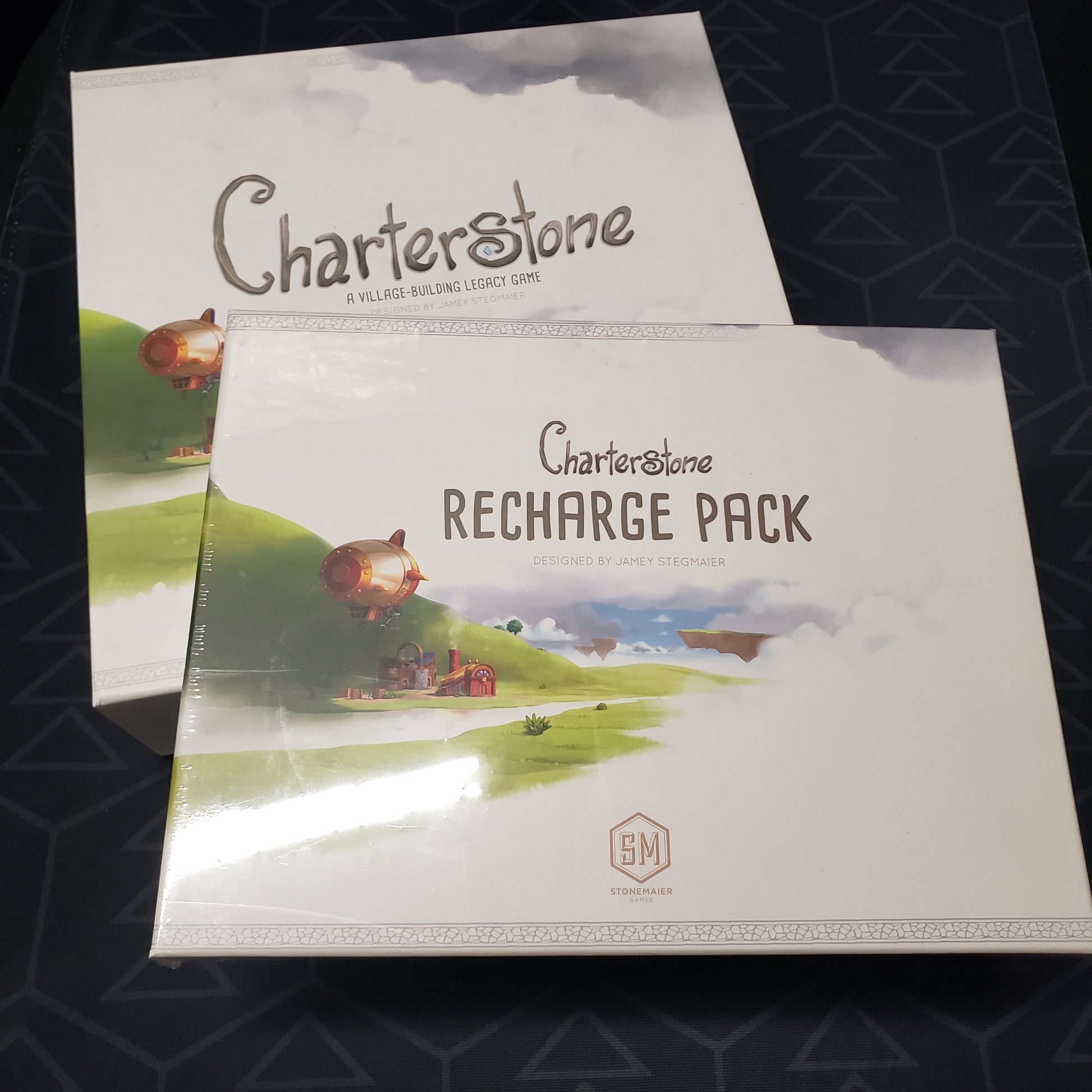 Image shows the front cover of the box for the Charterstone board game with the Recharge Pack for the game sitting on top of it