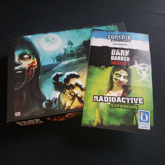 Image shows the front cover of the box of the Dark Darker Darkest board game, with the instructions for the Zombie and Radioactive expansions sitting on top of it