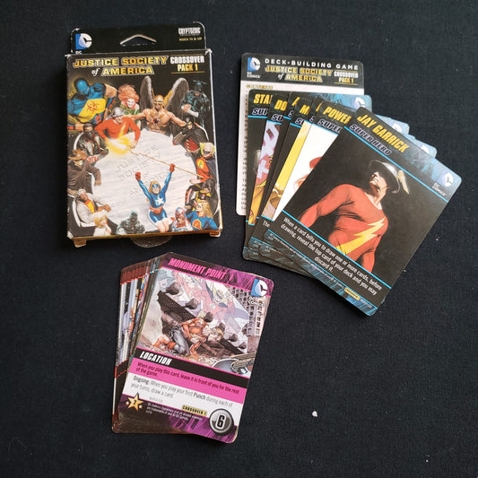 Image shows the front cover of the box of the Justice Society of America Crossover Pack expansion for the DC Comics Deckbuilding card game, along with the cards fanned out next to it