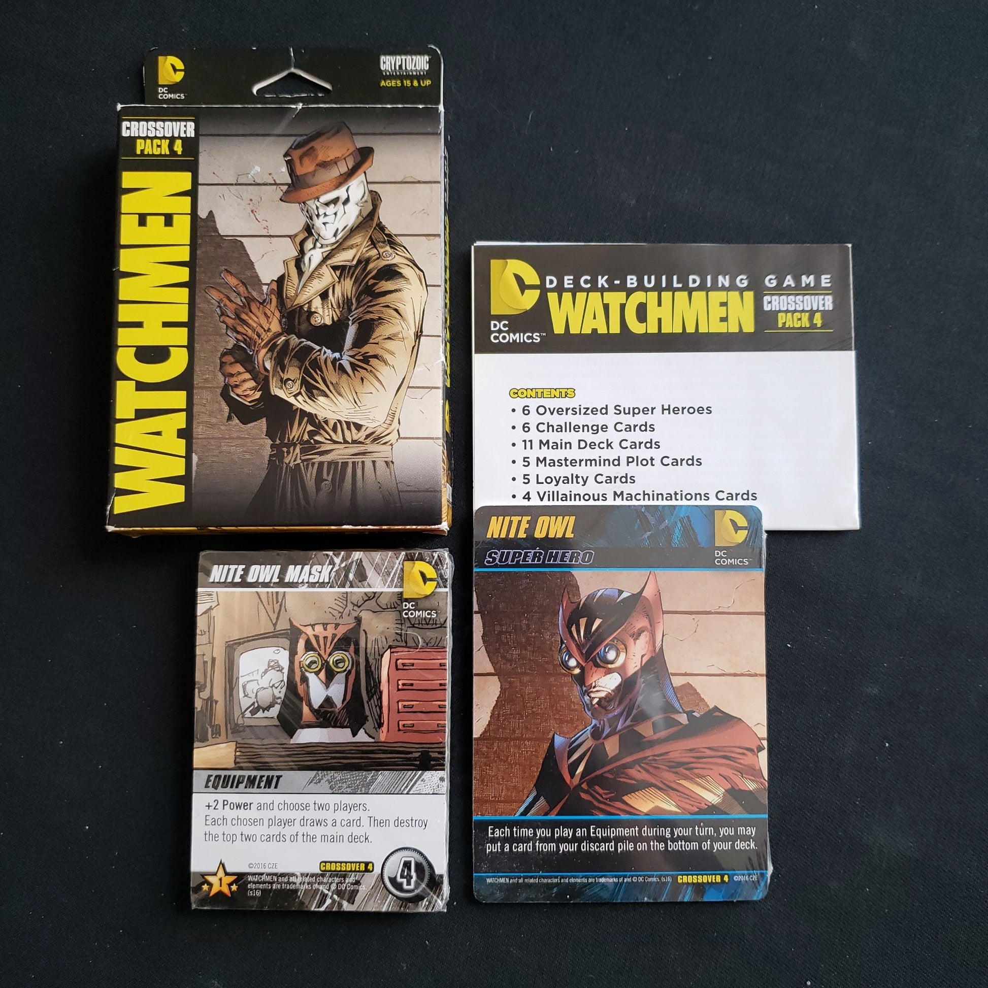 Image shows the front cover of the box of the Watchmen Crossover Pack expansion for the DC Comics Deckbuilding card game, along with the cards in shrinkwrap next to it