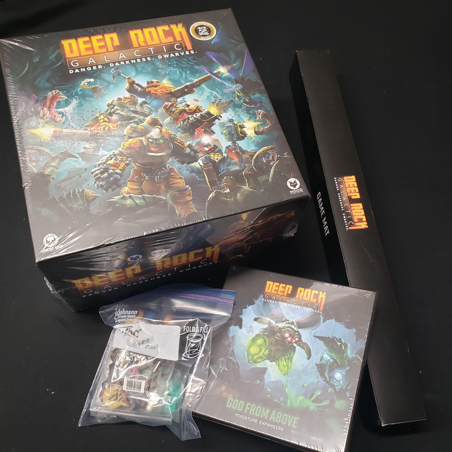 Image shows the front cover of the box for Deep Rock Galactic: The Board Game, the boxed game mat, expansion box and a baggie of extras sitting next to it