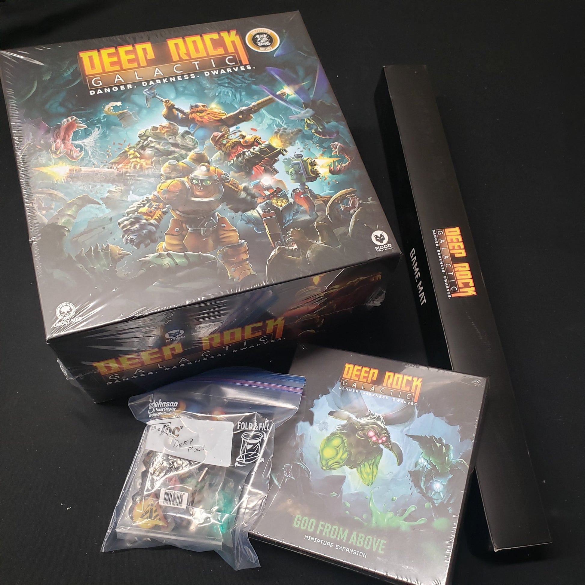 Image shows the front cover of the box for Deep Rock Galactic: The Board Game, the boxed game mat, expansion box and a baggie of extras sitting next to it