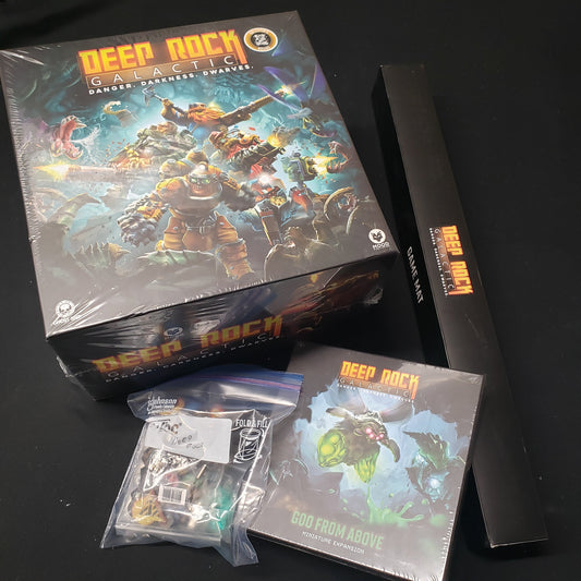 Image shows the front cover of the box for Deep Rock Galactic: The Board Game, the boxed game mat, expansion box and a baggie of extras sitting next to it