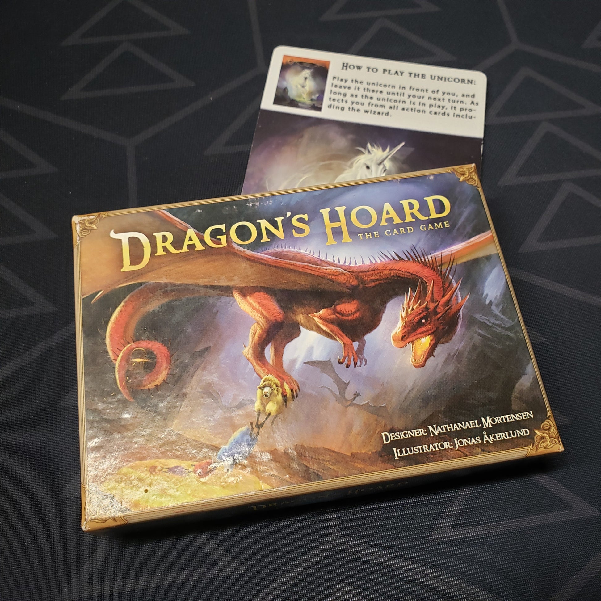 Image shows the front cover of the box for the Dragon's Hoard (First Edition) card game with the Unicorn promo card sitting underneath it
