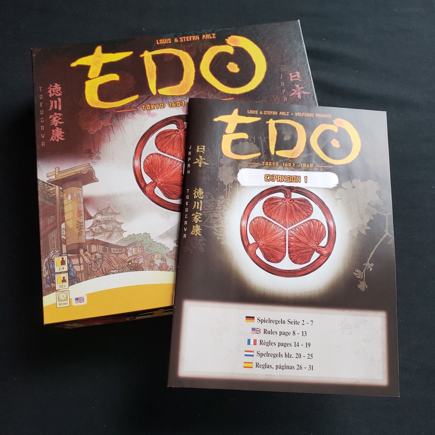 Image shows the front cover of the box of the Edo board game, with the instructions for Expansion #1 sitting on top of it