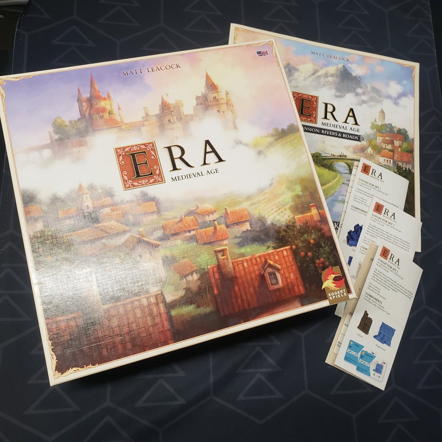 Image shows the front cover of the box for the Era: Medieval Age board game along with several expansion manuals