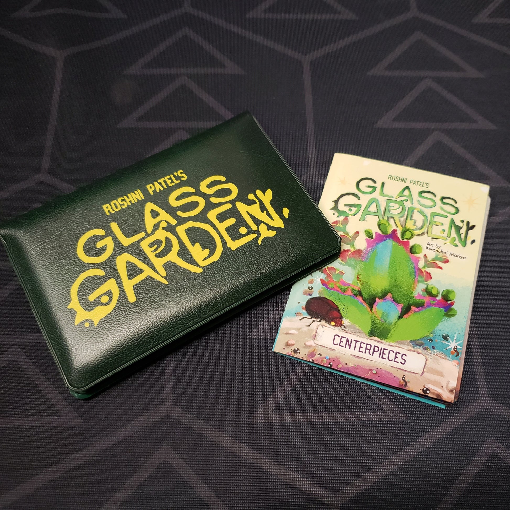 Image shows the front of the card wallet for the Glass Garden card game with the instructions for the Centerpieces expansion sitting next to it