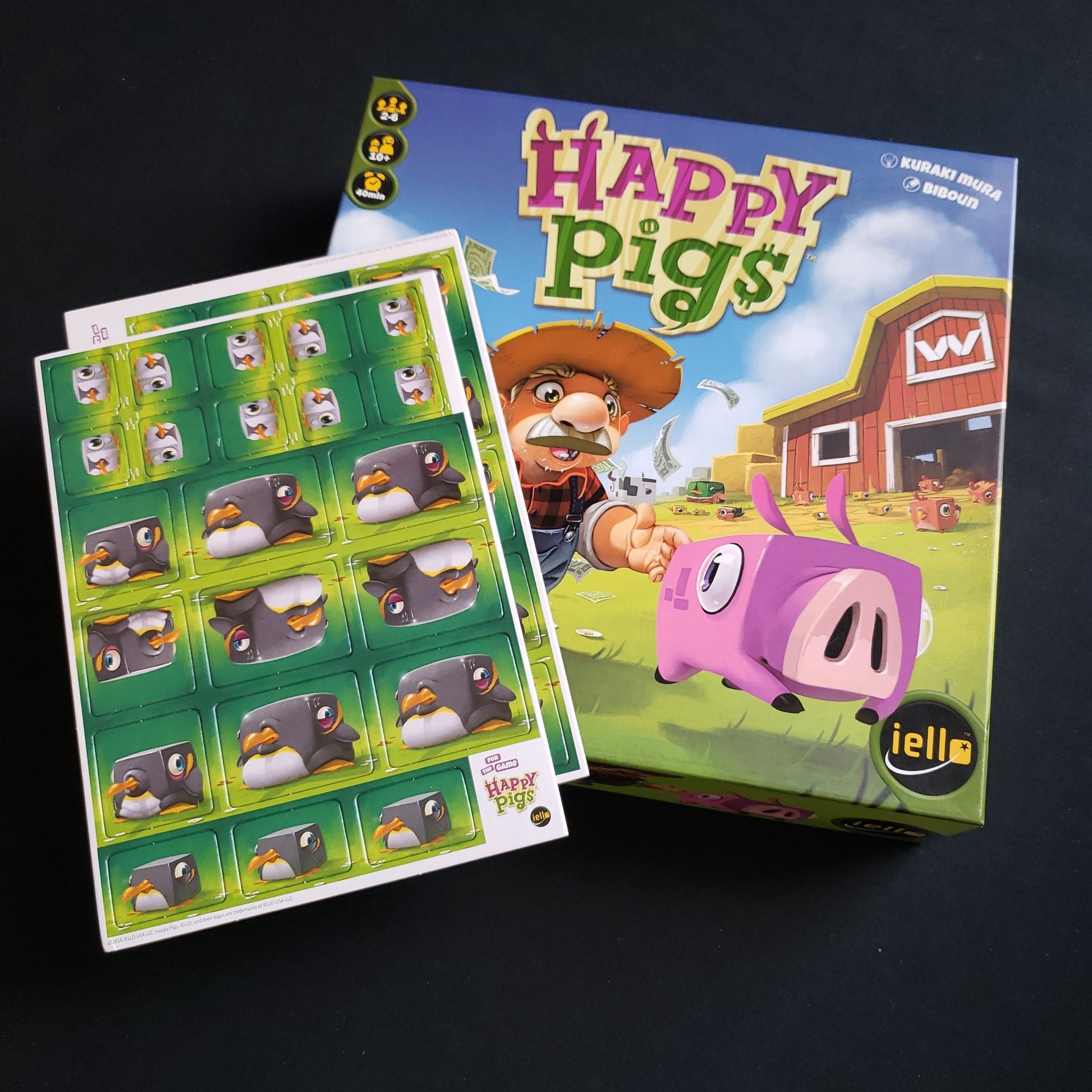 Image shows the front cover of the box of the Happy Pigs board game, with the Peguins promo tiles sitting on top of it