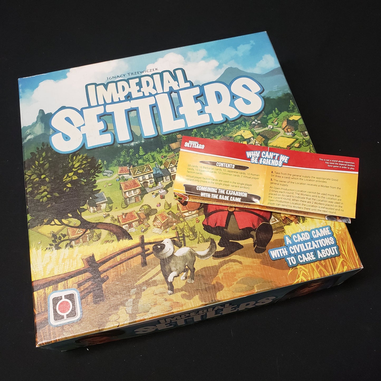 Image shows the front cover of the box of the Imperial Settlers card game with the instructions for the Why Can't We Be Friends expansion sitting on top of it