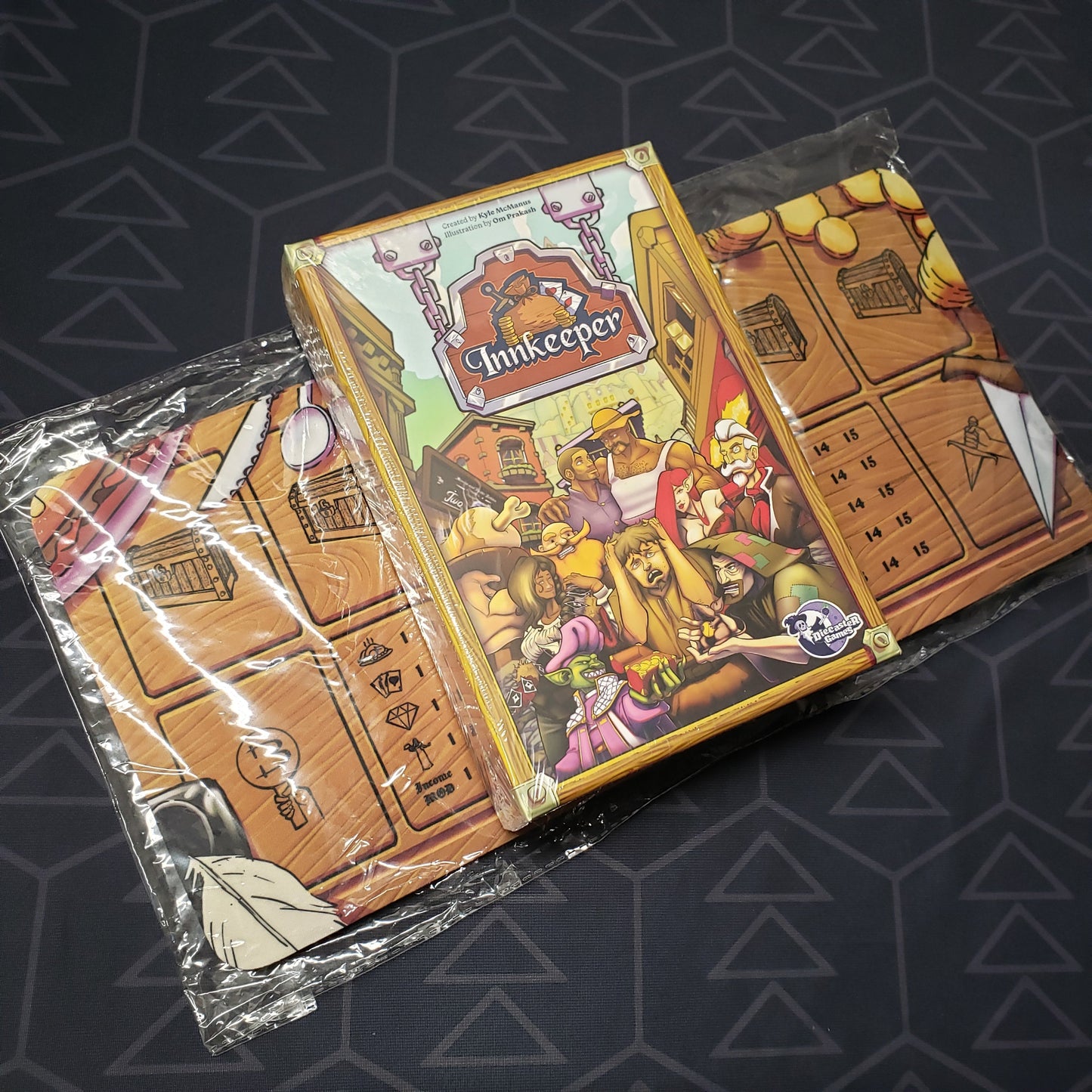 Image shows the front cover of the box for the Innkeeper card game sitting on top of a pack of game mats
