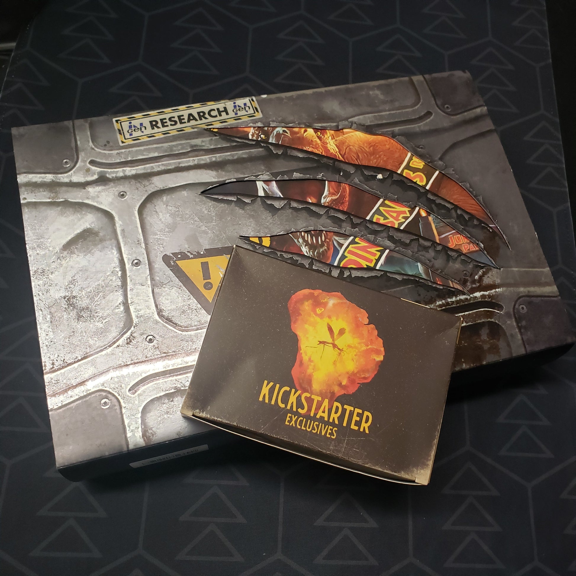 Image shows the front cover of the box for the Jurassic World: The Legacy of Isla Nublar board game with the Kickstarter Exclusives box sitting on top of it