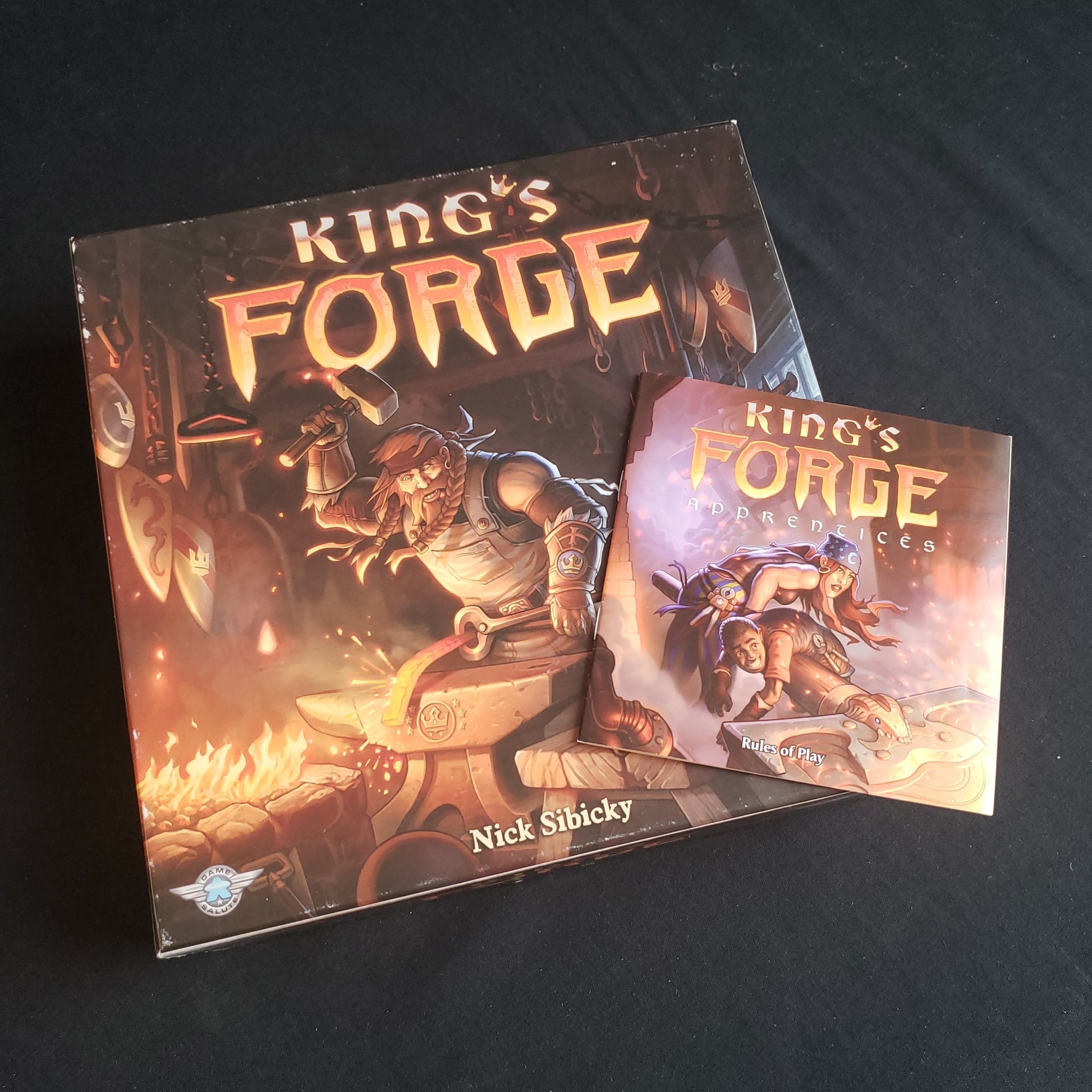 Image shows the front cover of the box of the King's Forge board game, with the instructions for the Apprentices expansion sitting on top of it