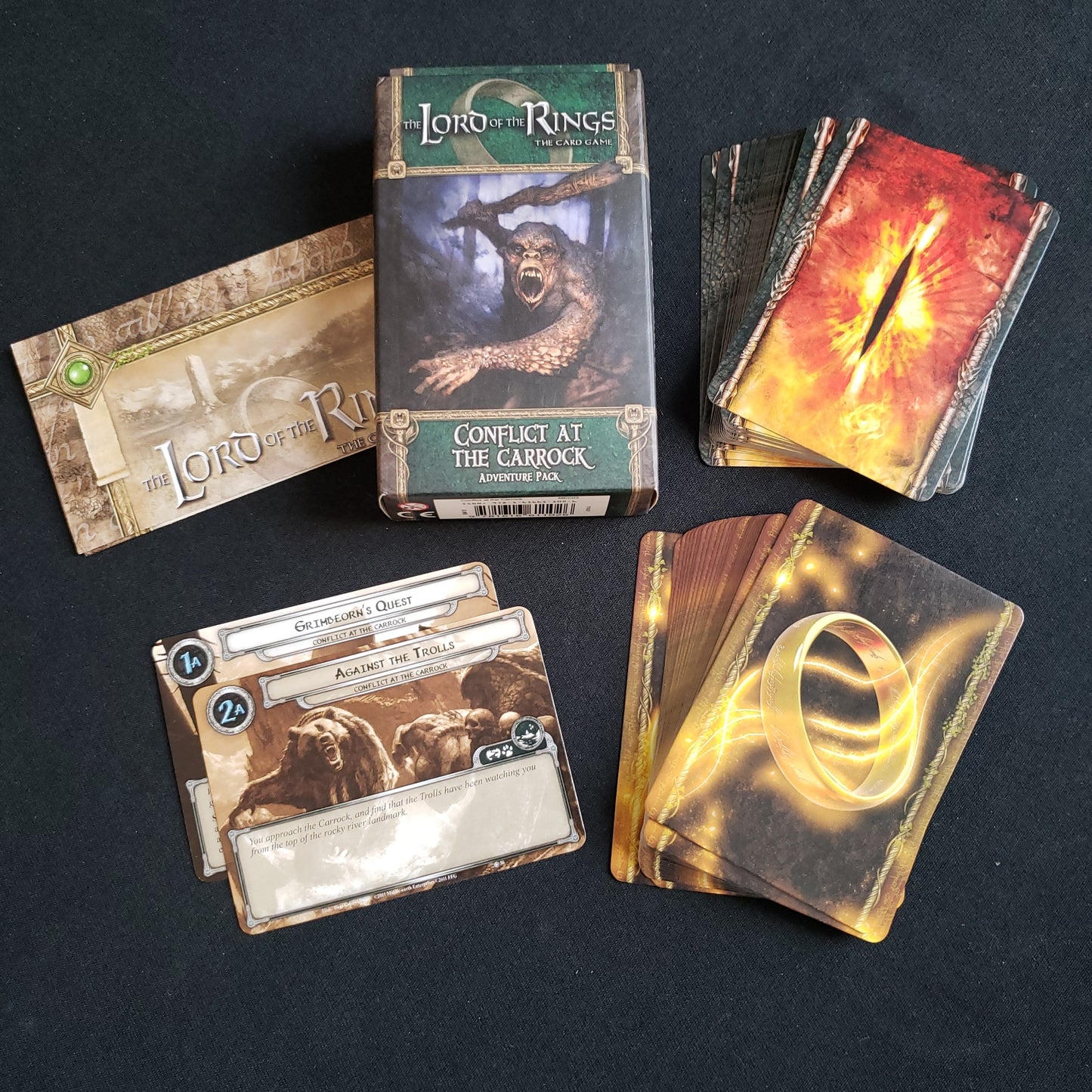 Image shows the front of the package for the Conflict at the Carrock Adventure Pack for the Lord of the Rings card game, with the cards and instructions fanned out around it