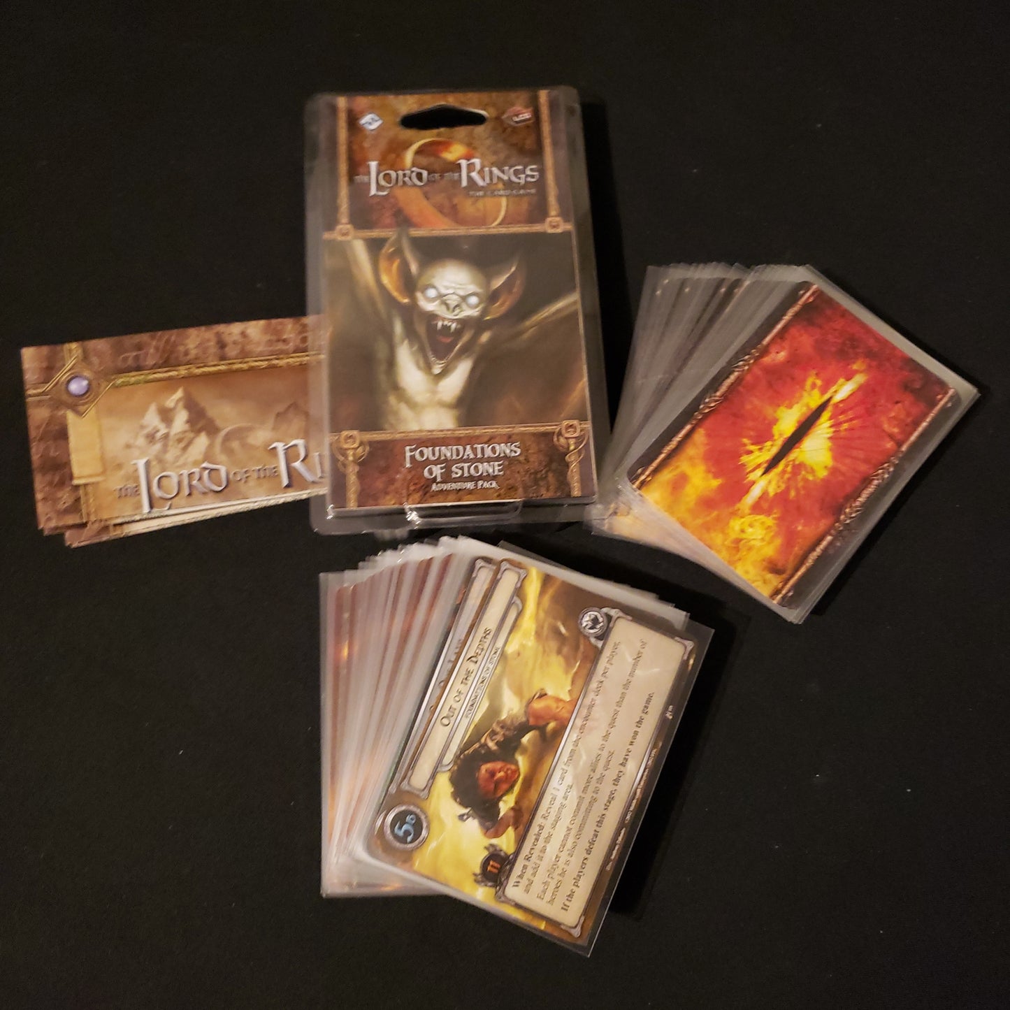 Image shows the front of the package for the Foundations of Stone Adventure Pack for the Lord of the Rings card game, with the cards and instructions fanned out around it