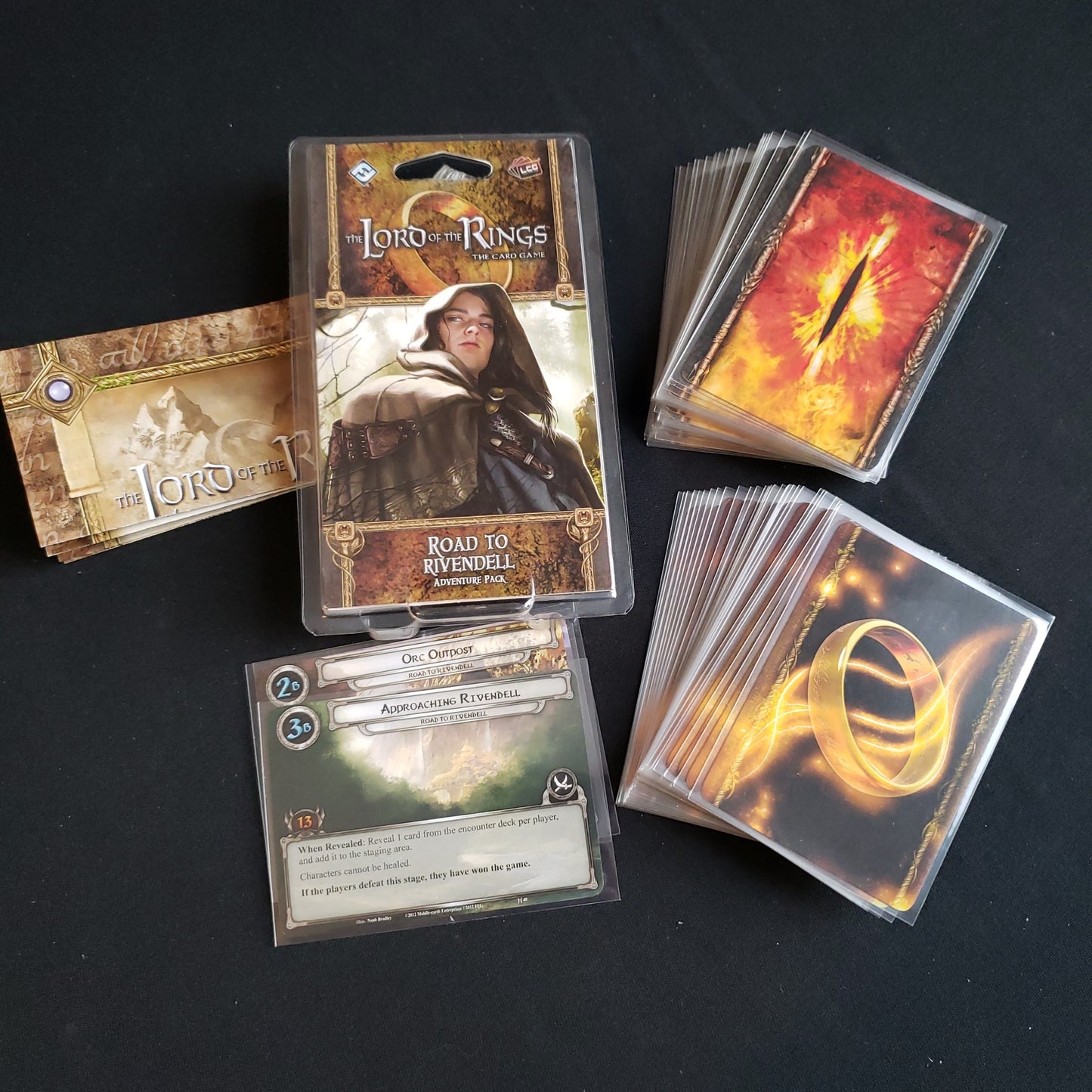 Image shows the front of the package for the Road to Rivendell Adventure Pack for the Lord of the Rings card game, with the cards and instructions fanned out around it