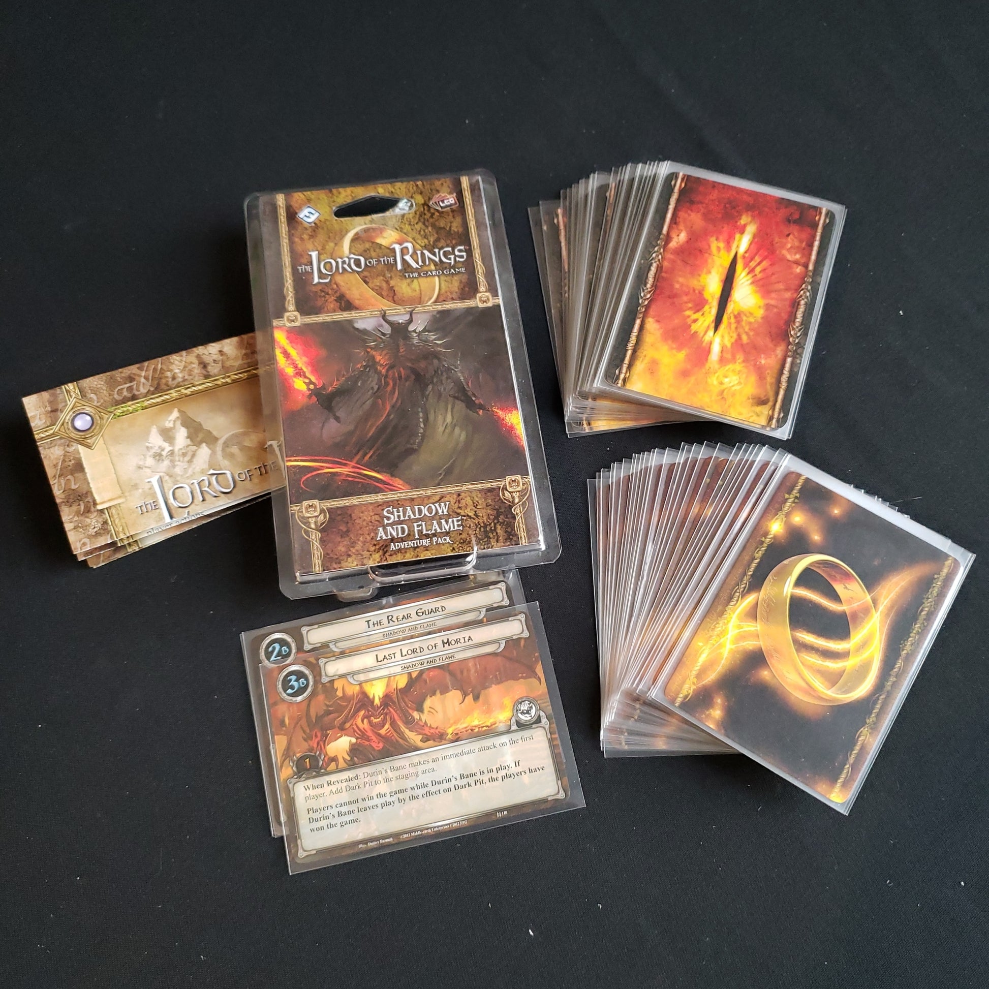 Image shows the front of the package for the Shadow and Flame Adventure Pack for the Lord of the Rings card game, with the cards and instructions fanned out around it