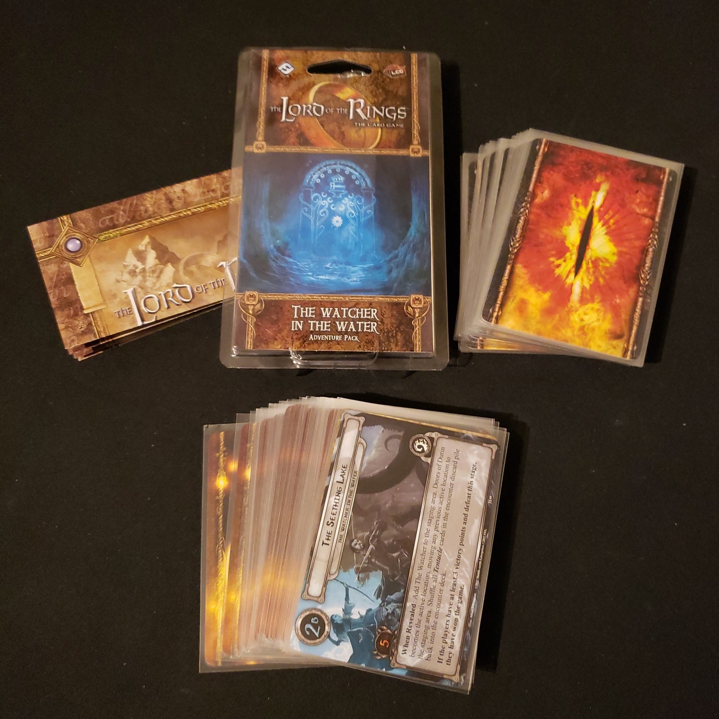 Image shows the front of the package for the Watcher in the Water Adventure Pack for the Lord of the Rings card game, with the cards and instructions fanned out around it