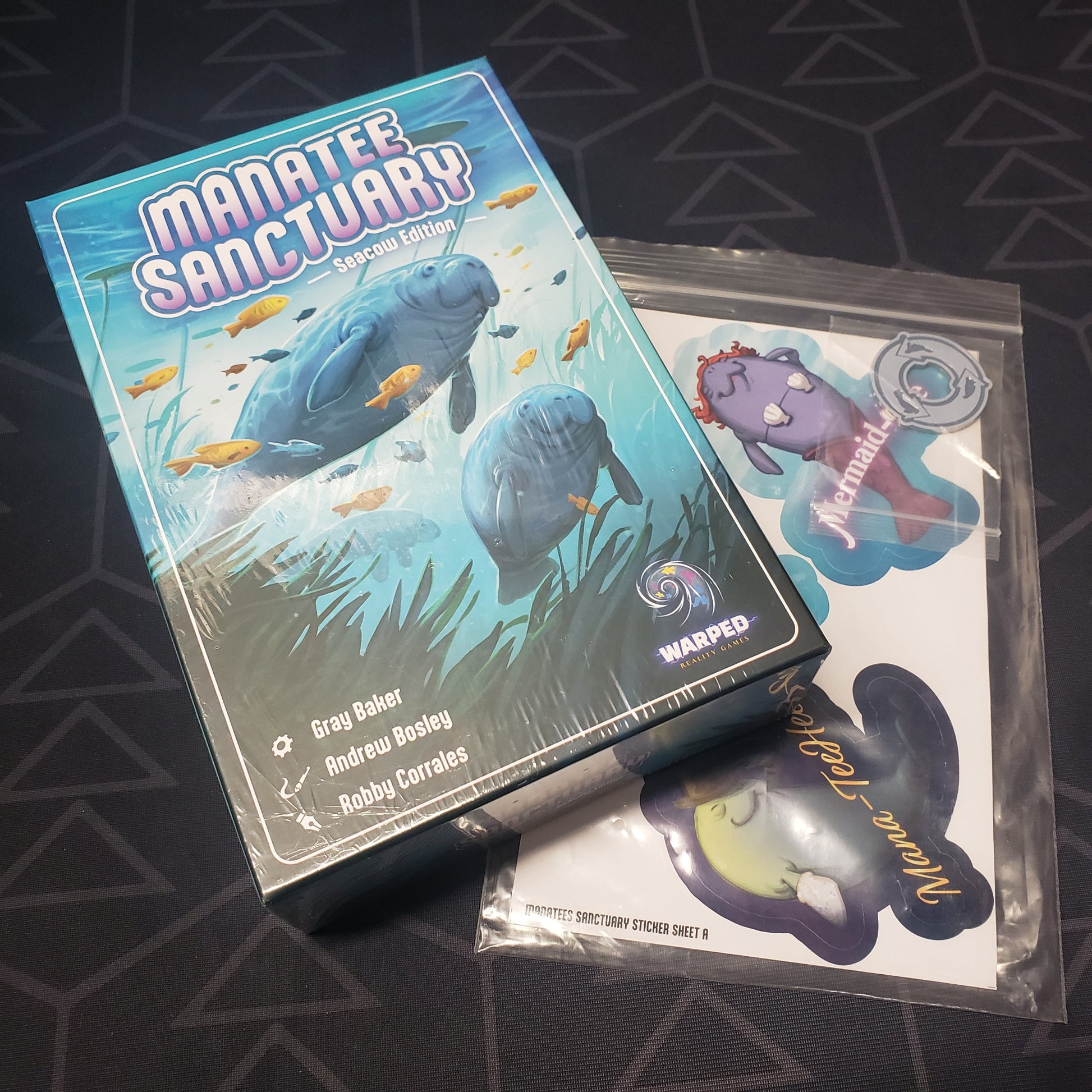 Image shows the front cover of the box for the Manatee Sanctuary (Seacow Edition) board game sitting on top of a bonus sticker pack