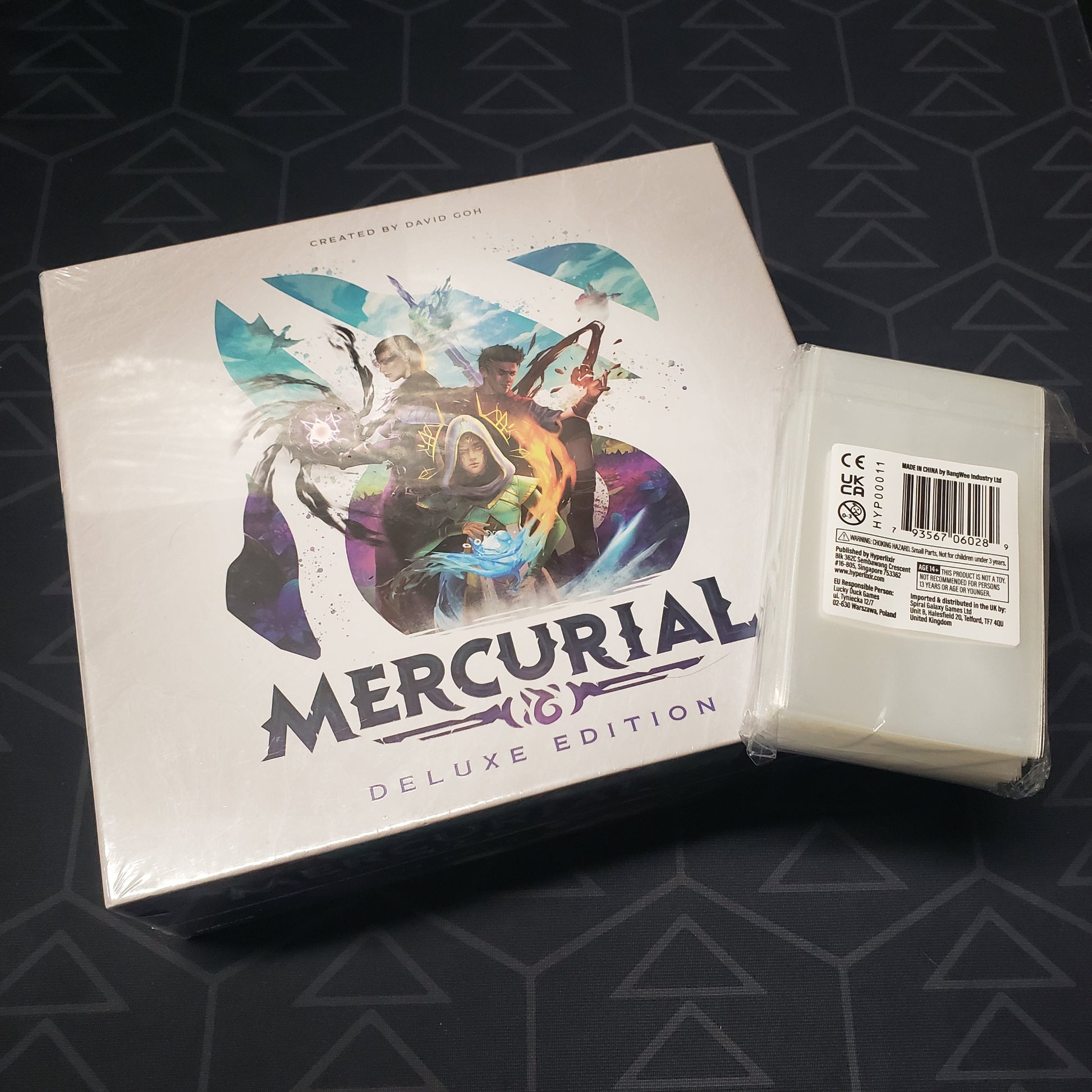 Image shows the front cover of the box for the Mercurial (Deluxe Edition) board game with a pack of clear card sleeves sitting on top of it