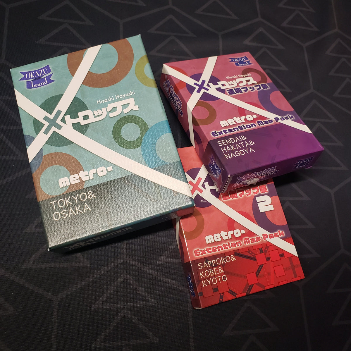 Image shows the front covers of the boxes for the Japanese / English printing of the board game MetroX, along with two of its expansions