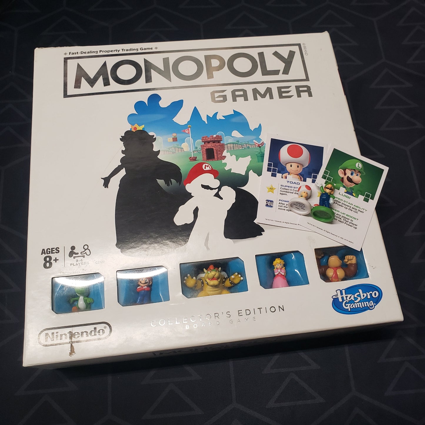 Image shows the front cover of the box for the Monopoly Gamer (Collector's Edition) board game with some bonus characters and their cards sitting on top of it