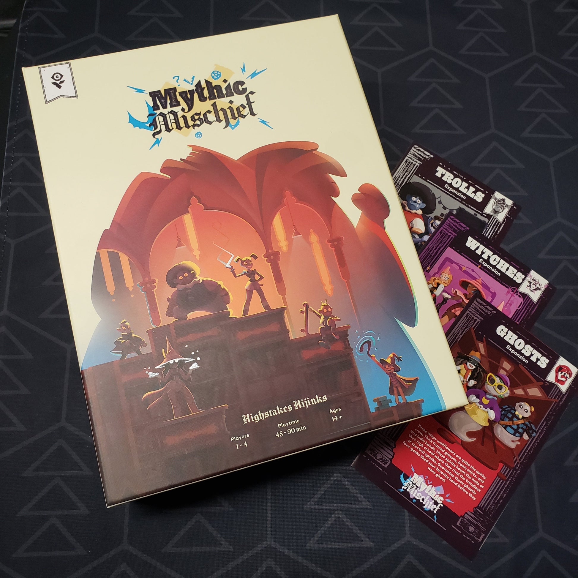 Image shows the front cover of the box for the Mythic Mischief board game sitting op top of the instruction sheets for three expansions