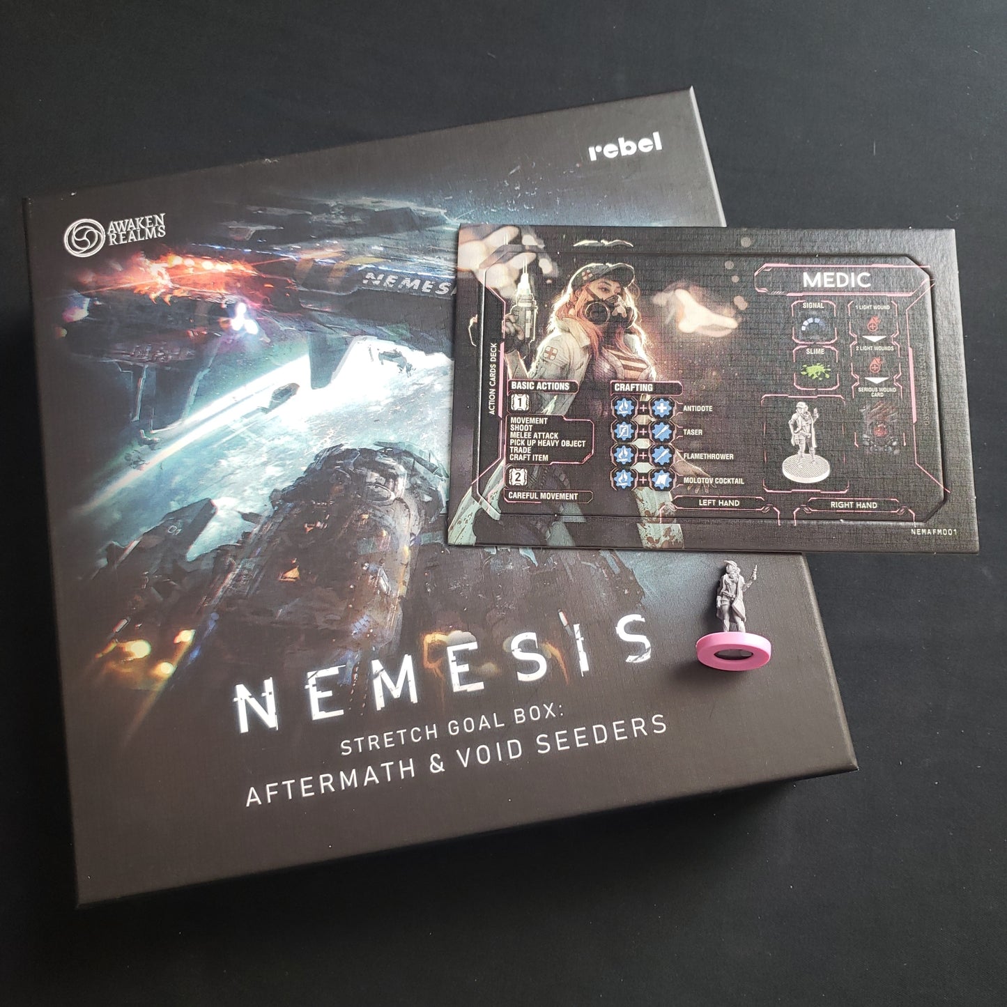 Image shows the front cover of the box of the Aftermath & Void Seeders Stretch Goals box for the board game Nemesis, with the character board and miniature for the Medic expansion sitting on top of it