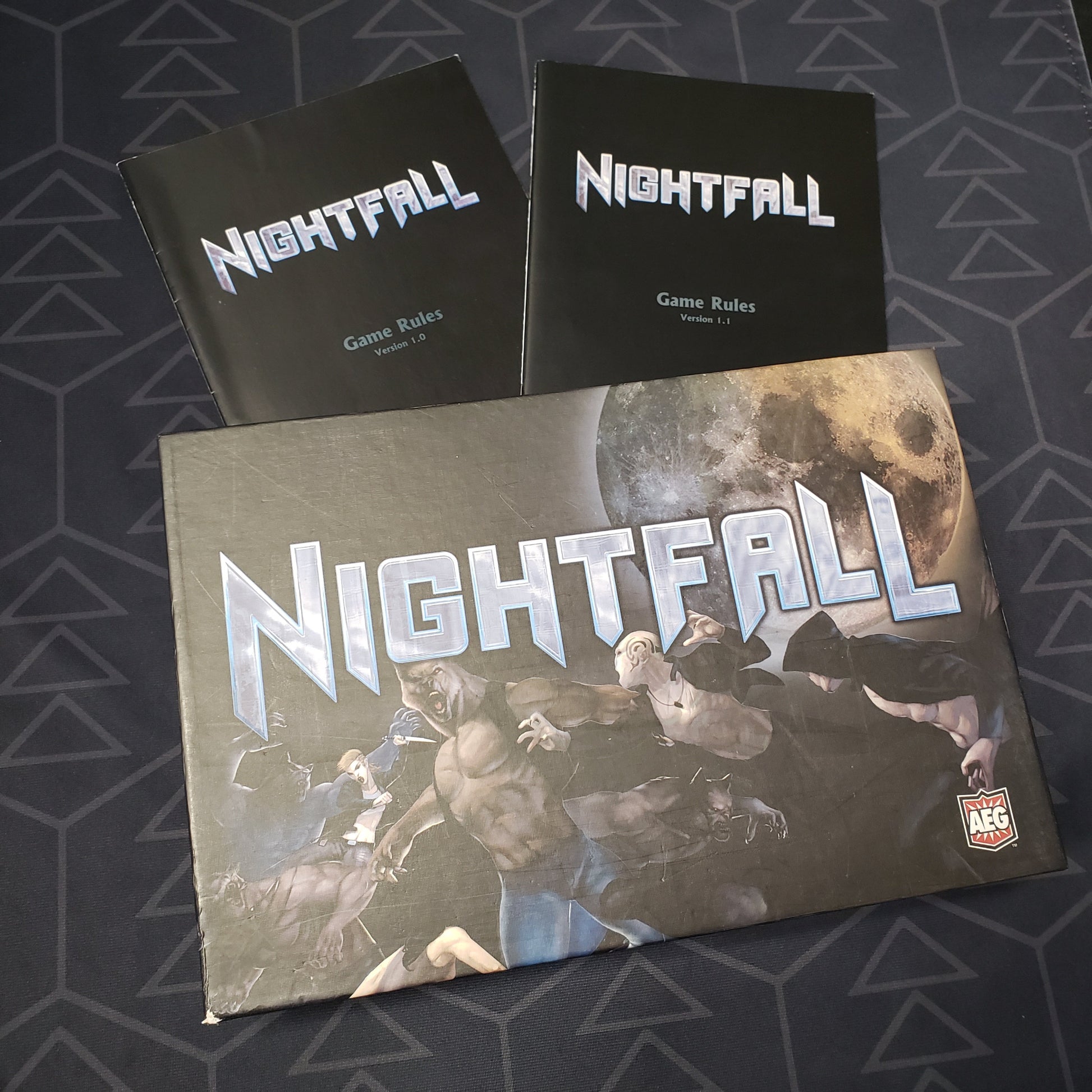 Image shows the front cover of the box of the Nightfall card game sitting on top of two instruction manuals