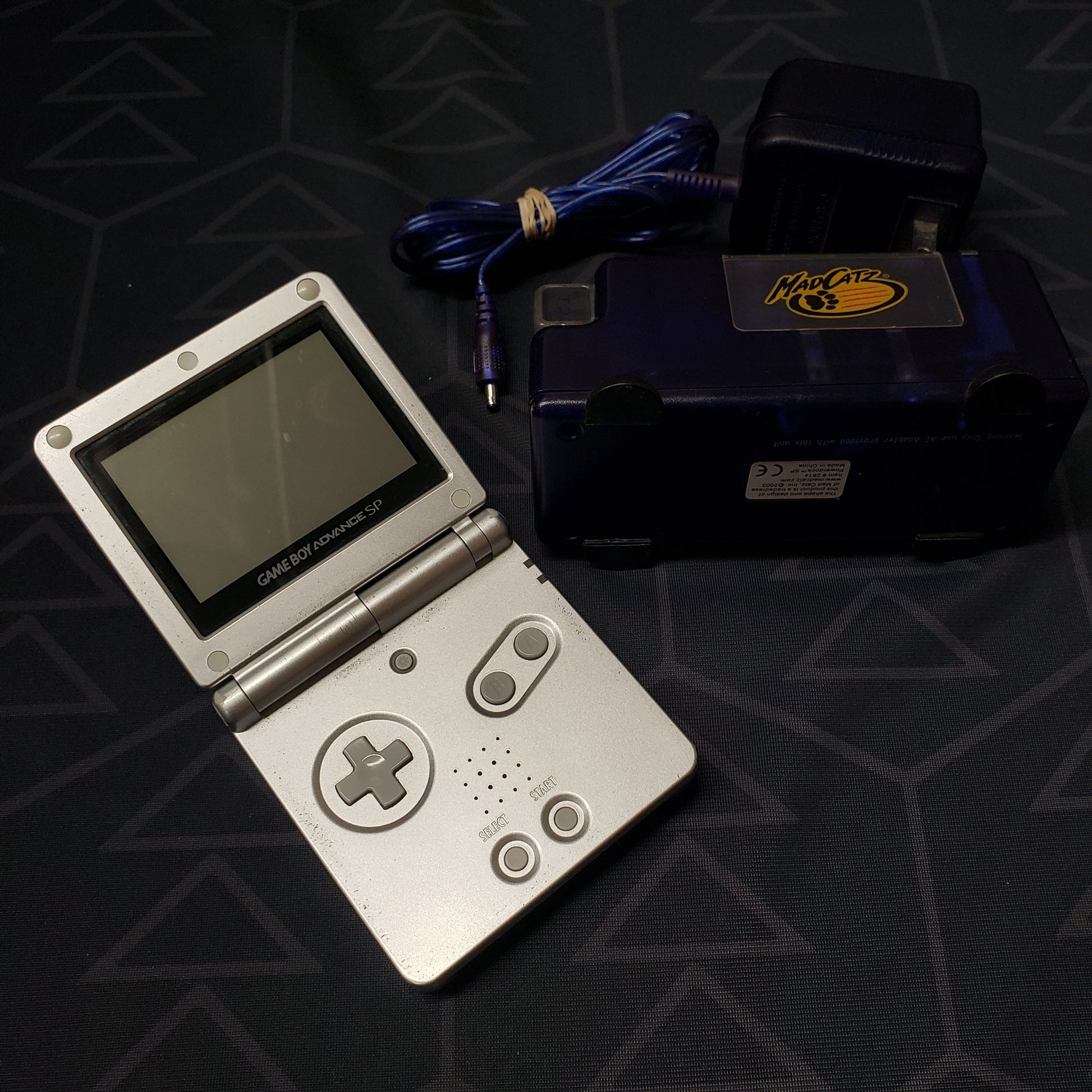 Image shows a platinum Nintendo Game Boy Advance SP gaming console sitting next to a Mad Catz charging station