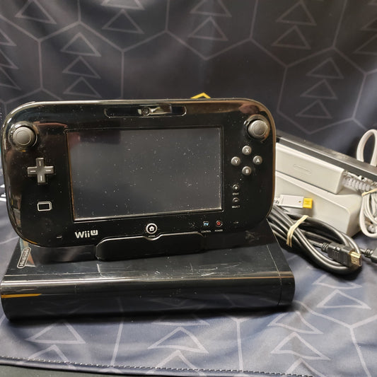 Image shows a black Nintendo Wii U console with its GamePad controller sitting in a stand on top of it & connecting cables wrapped in a pile