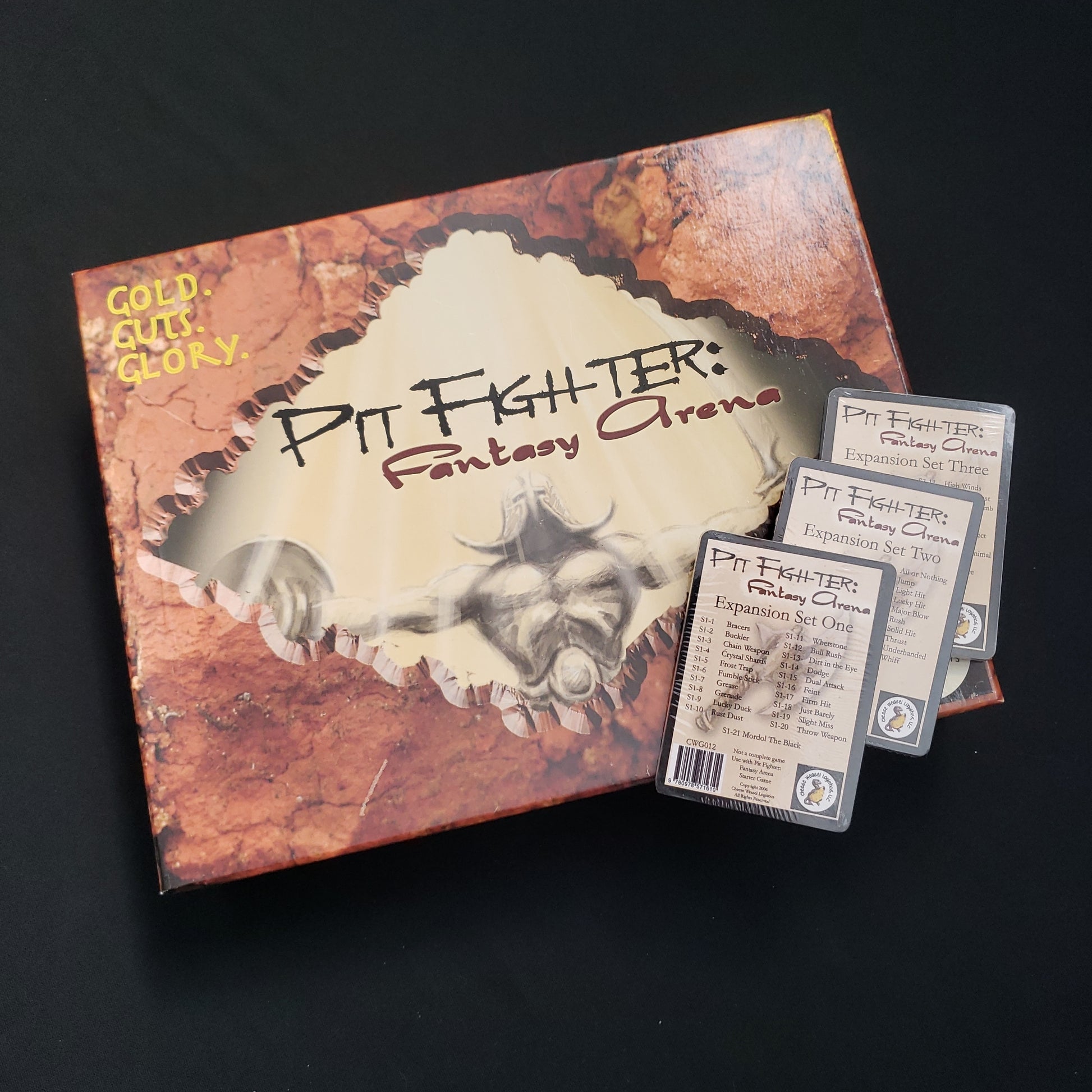 Image shows the front cover of the box of the Pit Fighter: Fantasy Arena board game, with  the sealed expansion decks 1, 2, and 3 sitting on top of it