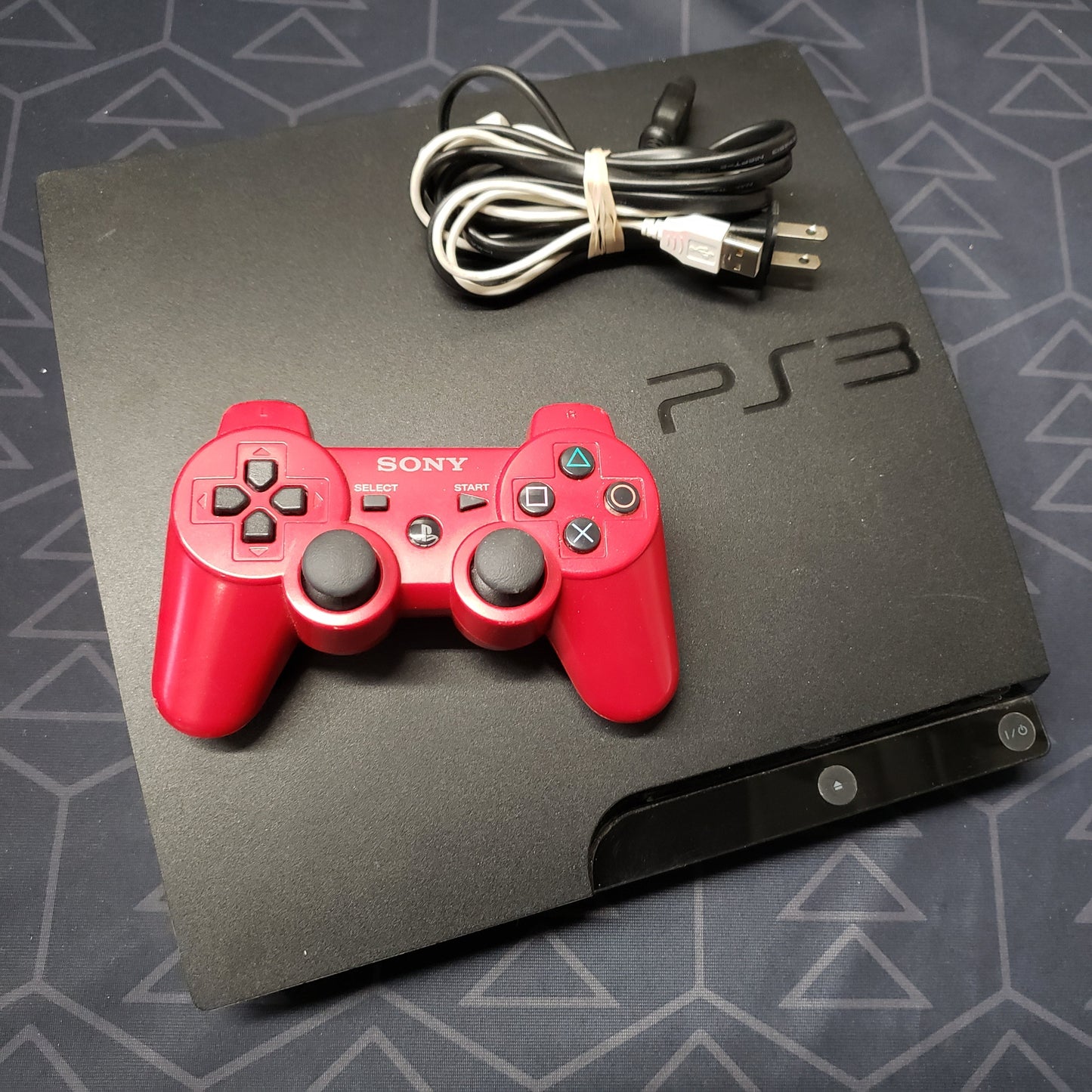 Image shows the top of a Playstation 3 Slim gaming console with a controller and cables sitting on top of it